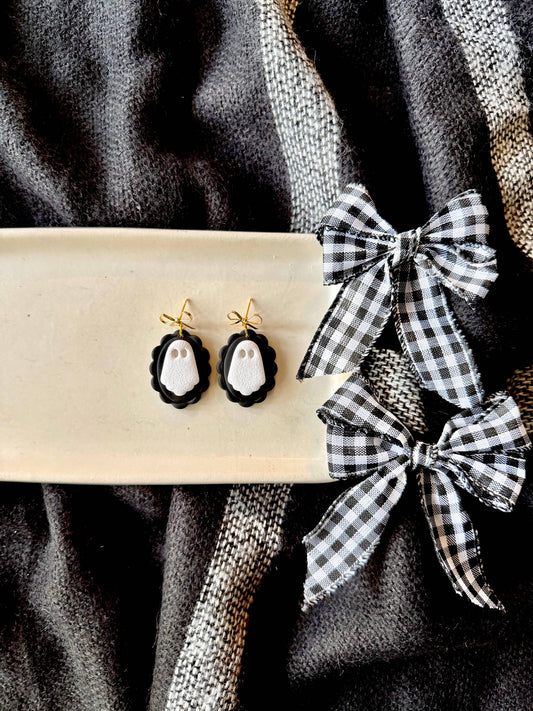 Black and white ghost-themed drop earrings displayed on a cream-colored rectangular tray, next to two black and white gingham ribbon bows on a dark textured fabric background.