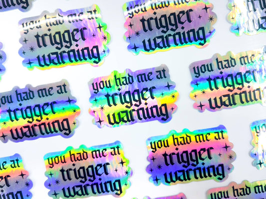 Holographic stickers with 'you had me at trigger warning' text on a white background