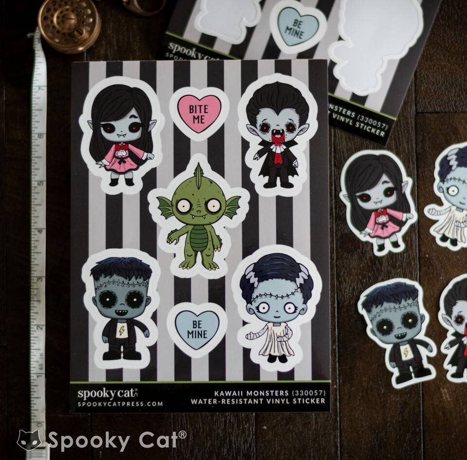 A sheet of Spooky Cat 'Kawaii Monsters' water-resistant vinyl stickers displayed on a dark wood surface next to a metal ruler. The sheet features cartoon monster characters including Frankenstein's monster, Dracula, and a green monster, alongside hearts that say 'BITE ME' and 'BE MINE'.