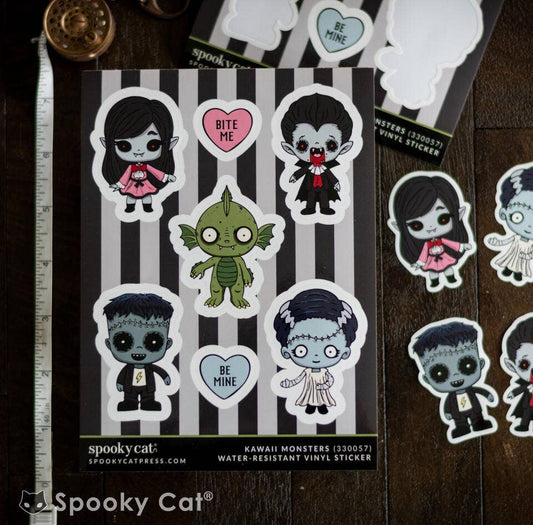 A sheet of Spooky Cat 'Kawaii Monsters' water-resistant vinyl stickers displayed on a dark wood surface next to a metal ruler. The sheet features cartoon monster characters including Frankenstein's monster, Dracula, and a green monster, alongside hearts that say 'BITE ME' and 'BE MINE'.
