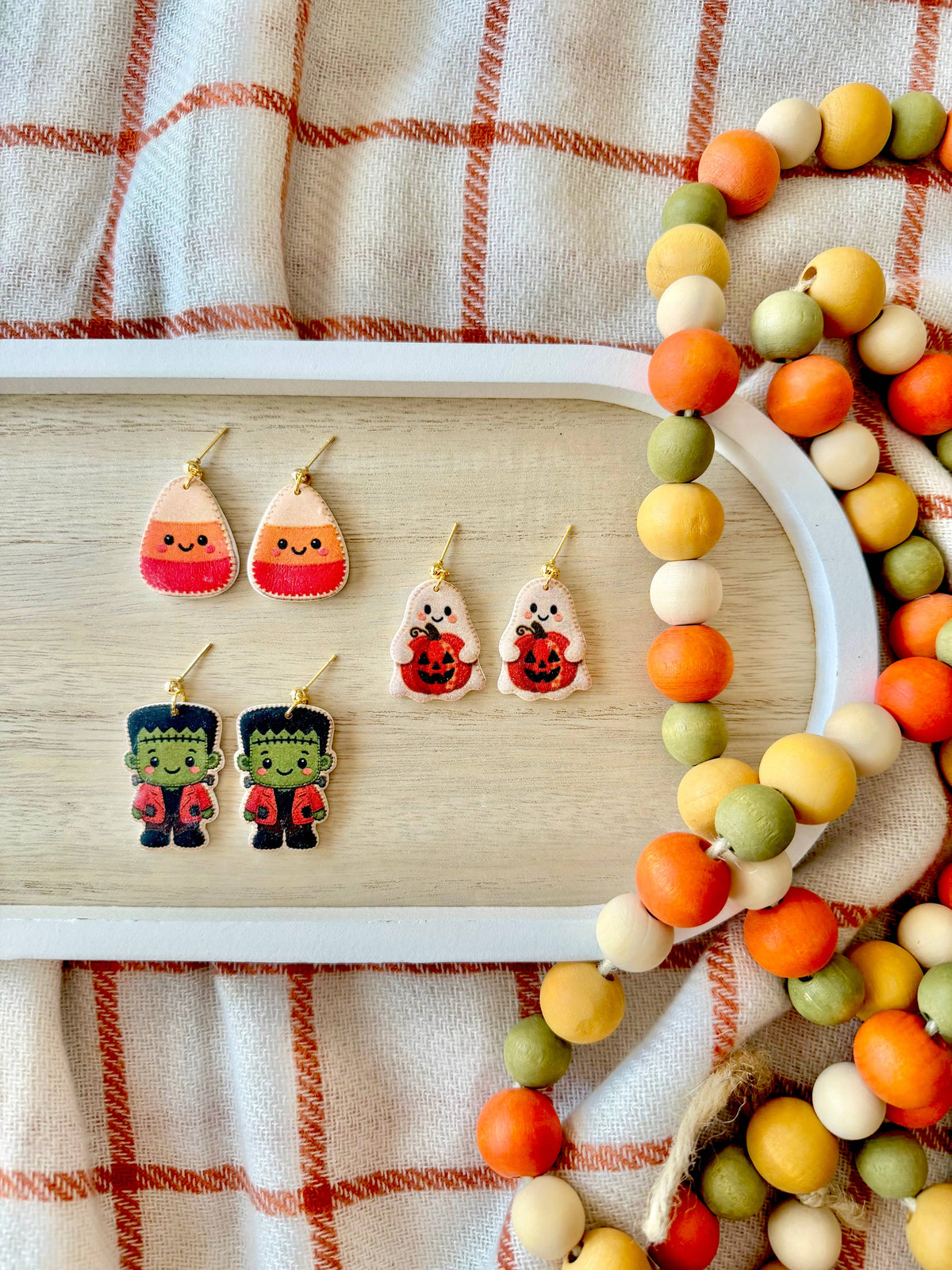 A styled flat-lay of Halloween-themed earrings displayed on a light wood tray, surrounded by a beaded garland on a cream plaid fabric background. The earrings include candy corn, jack-o'-lantern, and Frankenstein's monster designs.