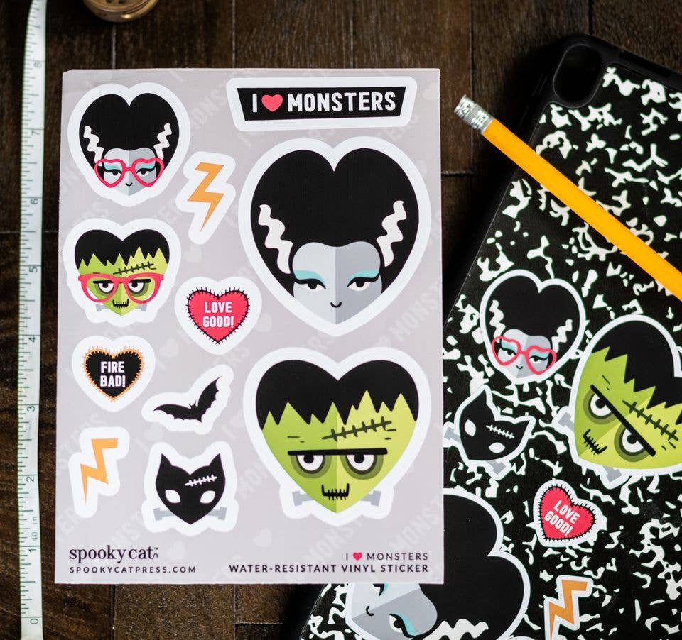 A sheet of Halloween-themed vinyl stickers featuring cartoon monsters, a cat, lightning bolts, and hearts, displayed on a dark wood surface next to a ruler and a black-and-white patterned phone case with matching stickers.