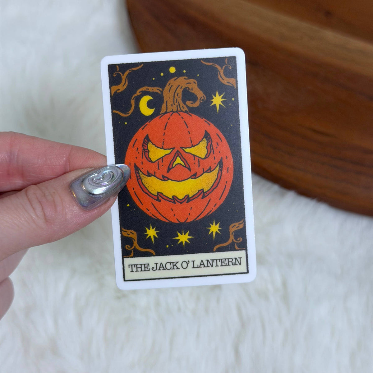 A hand with silver nail polish holds a tarot card featuring 'The Jack O' Lantern' illustration against a black background, with a white faux fur surface and a wooden bowl in the background.