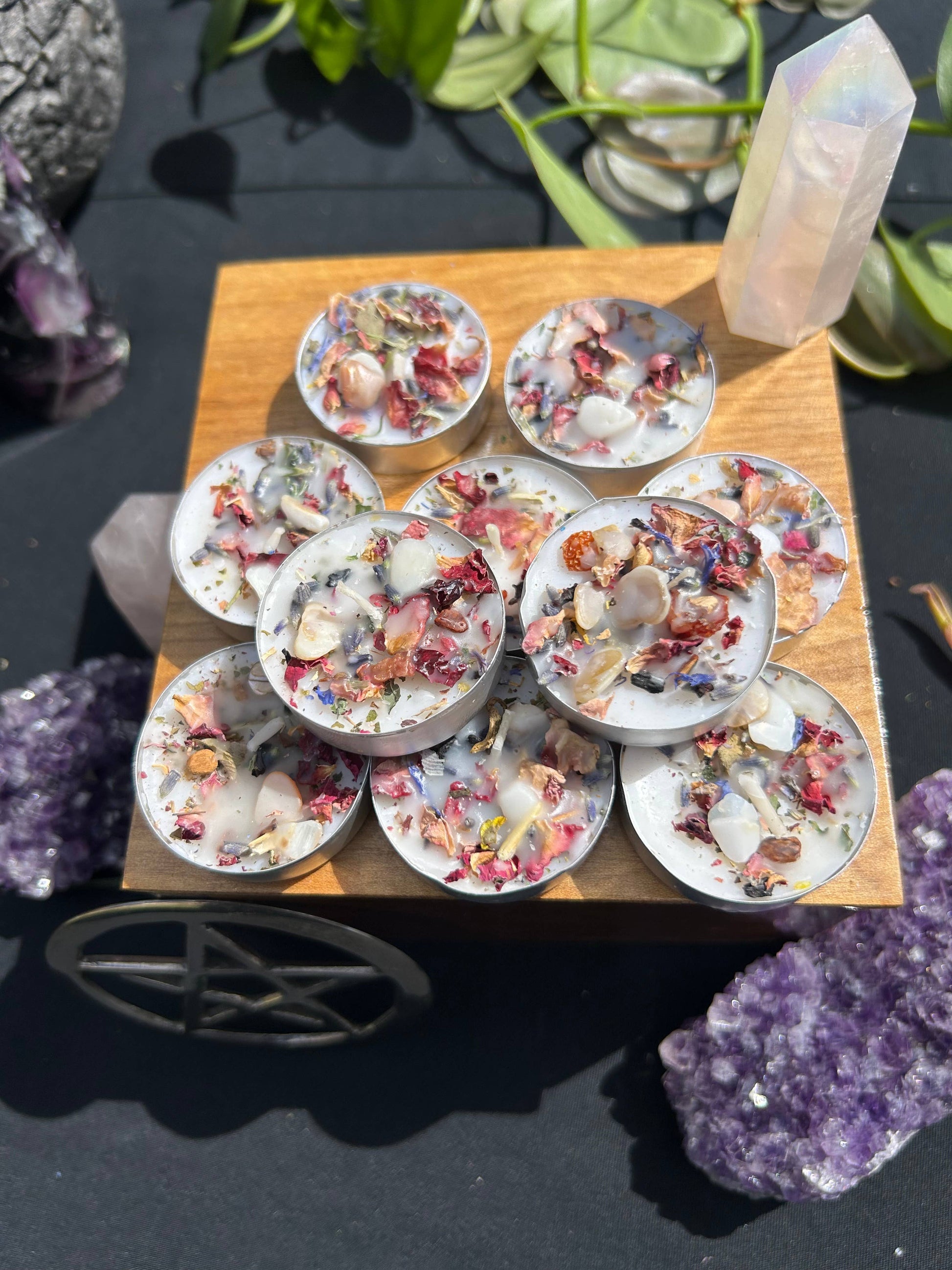 Small candles with herbs on a wooden board, surrounded by crystals and plants.