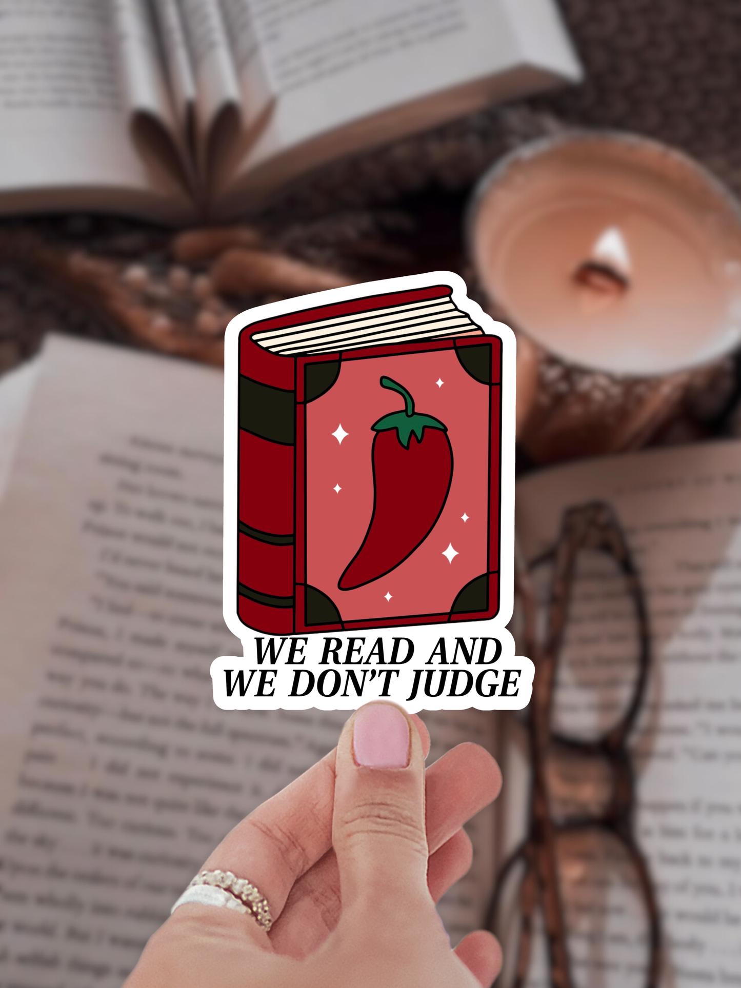 Sticker of a book with a chili pepper and 'We Read and Don't Judge' text held by a hand over an open book.