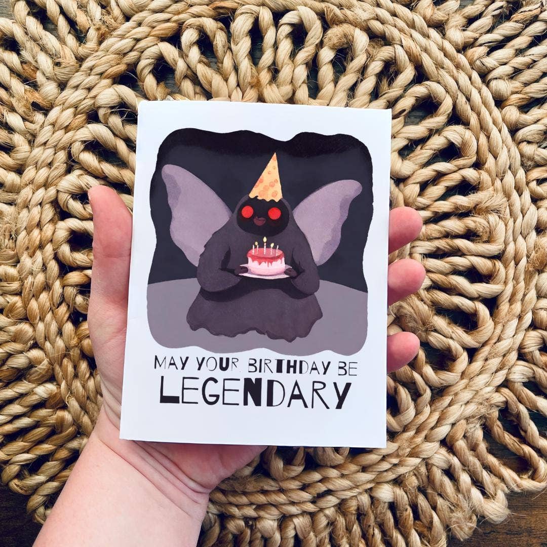 Hand holding a card with a Mothman holding a cake and text 'May your birthday be legendary' on a woven mat background.