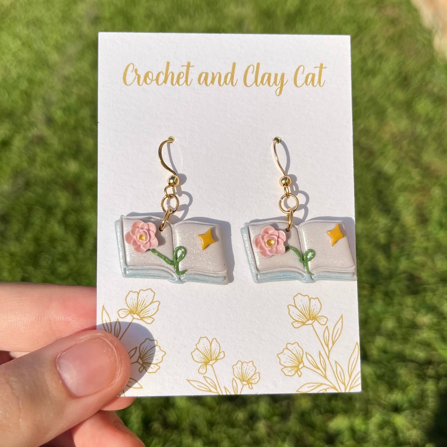 Pair of earrings on a 'Crochet and Clay Cat' branded card held by a hand with a grassy background.