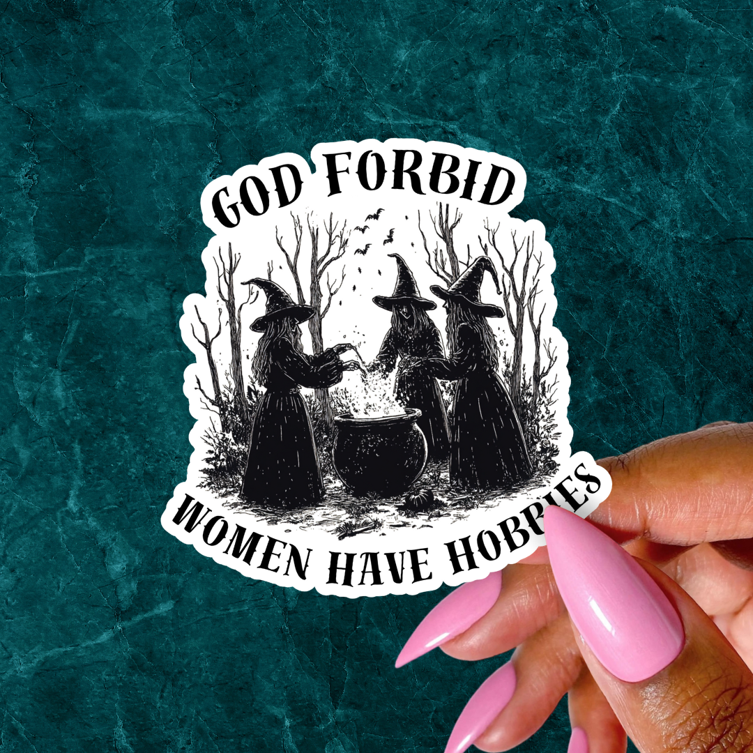 Sticker with witches and text 'God Forbid Women Have Hobbies' held by a hand with pink nails on a textured teal background.