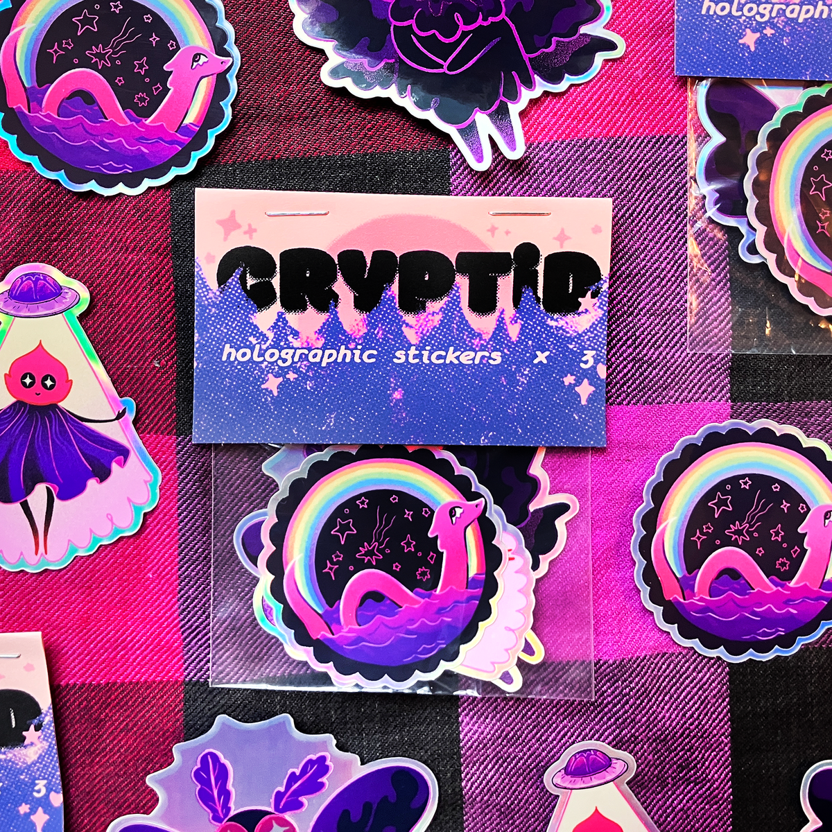 A set of colorful holographic stickers featuring a purple snake in a starry night sky under a rainbow, displayed on a pink and black checkered background. A central card reads 'CRYPTIC holographic stickers'.