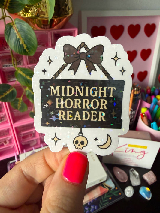 A hand with bright pink nail polish holds a die-cut sticker that reads 'MIDNIGHT HORROR READER' in gold lettering on a black background, decorated with a brown bow, stars, a skull, and a crescent moon. The background is a colorful, cluttered desk with pink storage organizers, crystals, and stationery.
