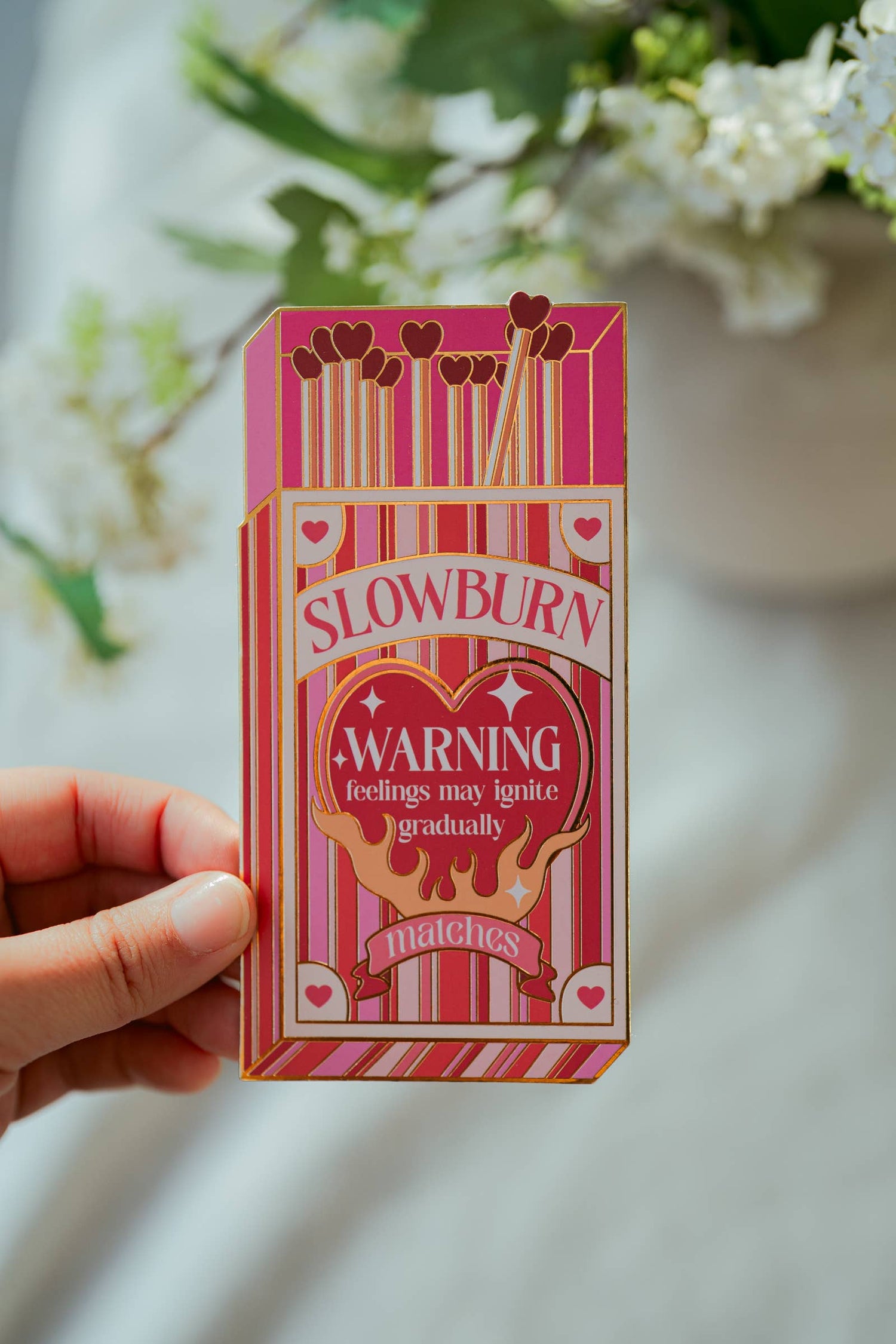 Pink matchbox-shaped bookmark labeled 'Slowburn Matches' held by a hand with a blurred floral background