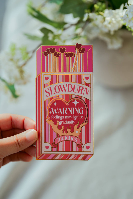 Pink matchbox-shaped bookmark labeled 'Slowburn Matches' held by a hand with a blurred floral background