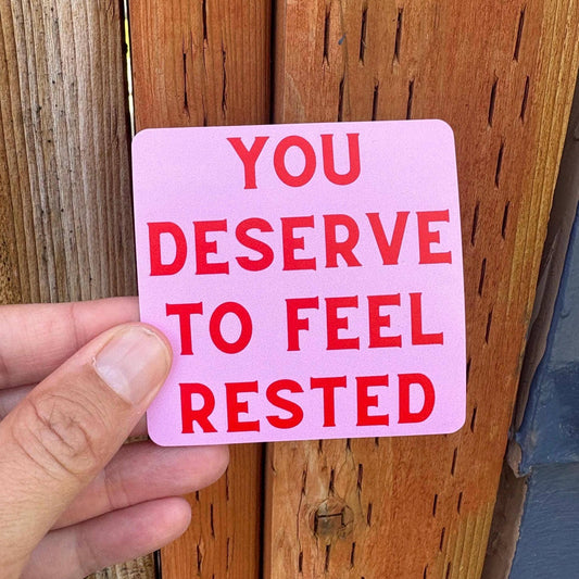 A hand holds a square pink sticker with bold red text that reads 'YOU DESERVE TO FEEL RESTED' against a wooden fence background.
