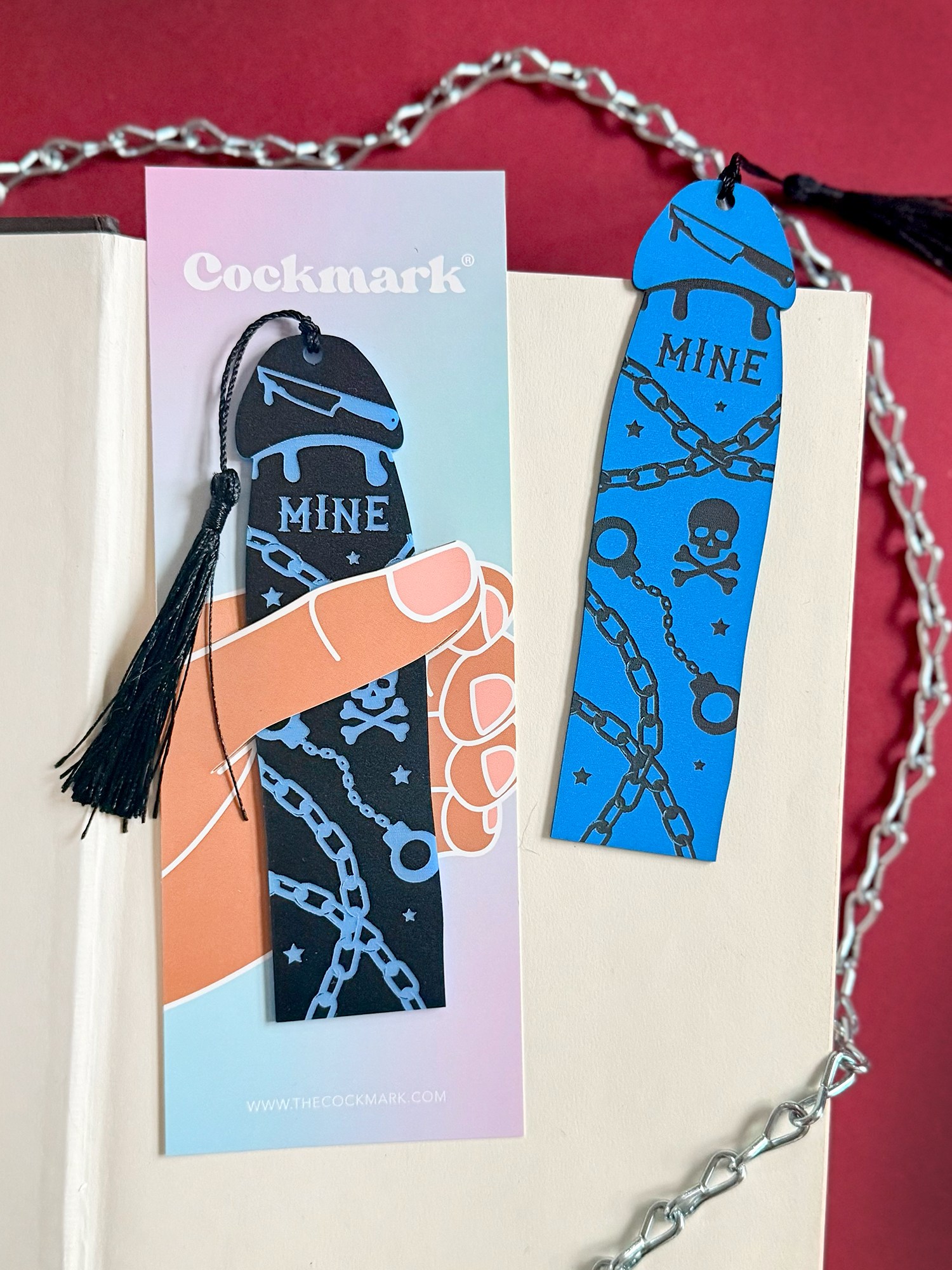 Blue and black 'Cockmark' bookmark with tassel on a pink card, against a red background.