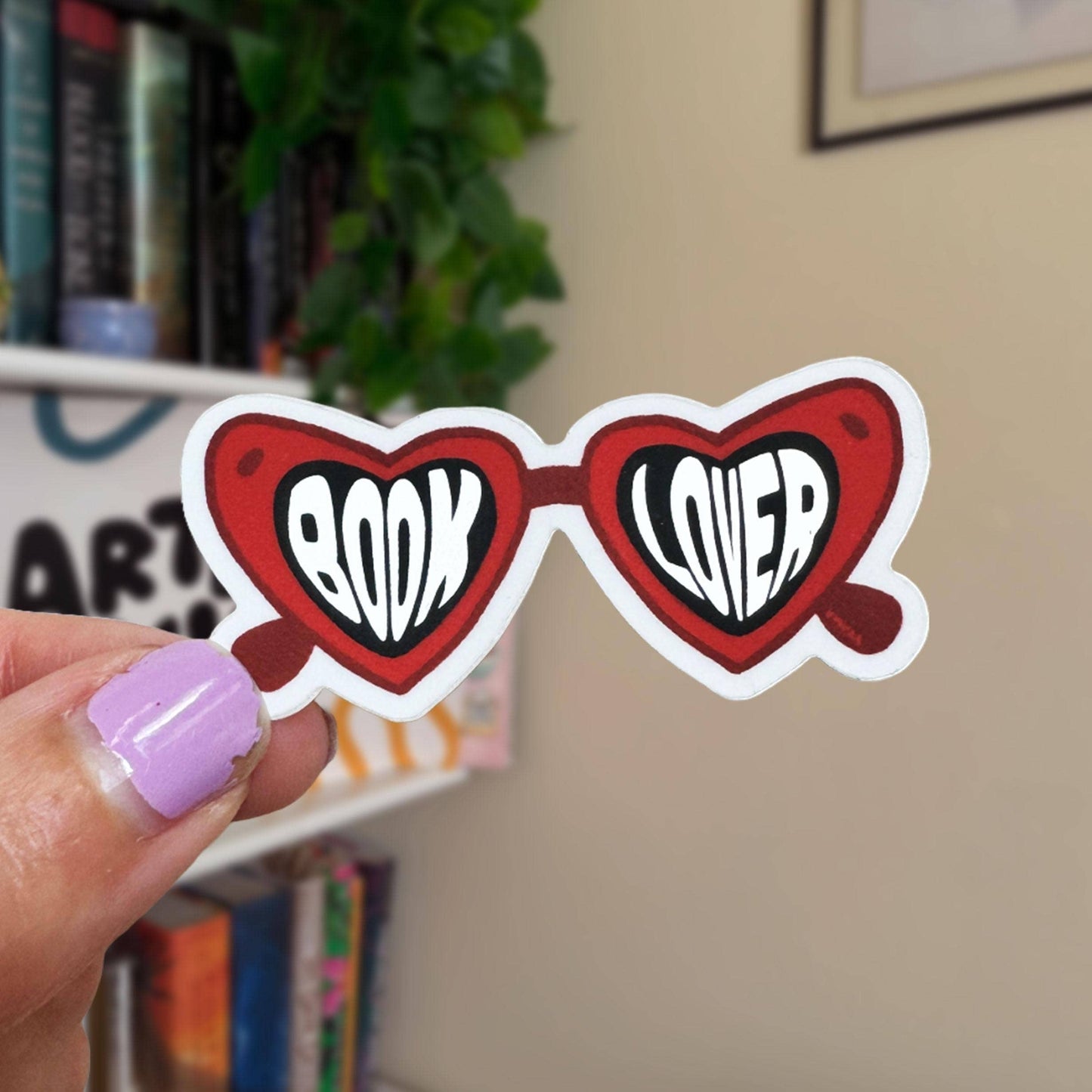 Heart-shaped sunglasses with 'BOOK LOVER' text held by a hand in front of a bookshelf.