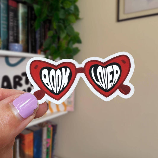 Heart-shaped sunglasses with 'BOOK LOVER' text held by a hand in front of a bookshelf.