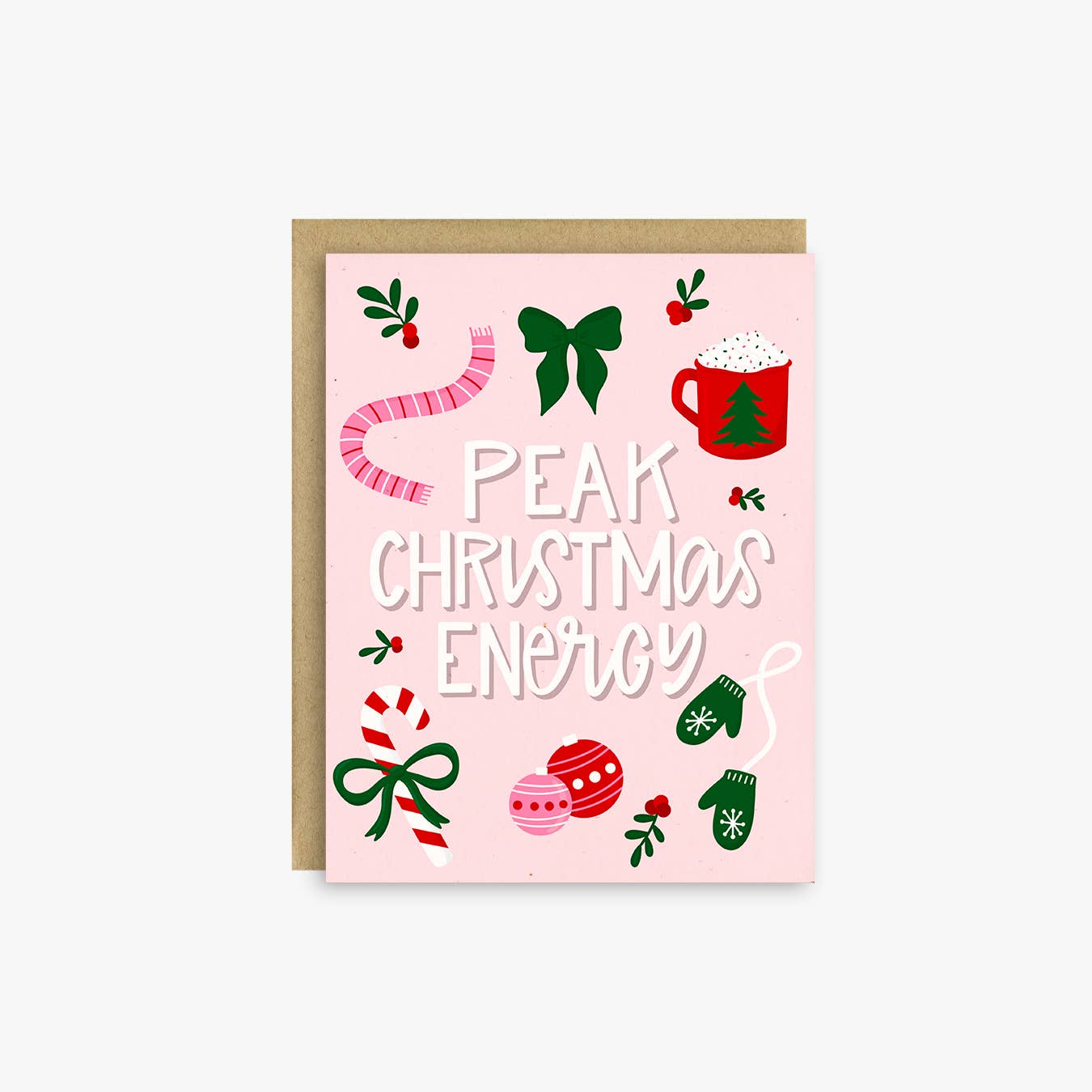 A pink Christmas greeting card with the phrase 'PEAK CHRISTMAS ENERGY' in white hand-drawn text, surrounded by festive illustrations including a green bow, red mug, candy cane, ornaments, and green mittens, shown with a kraft envelope on a light gray background.