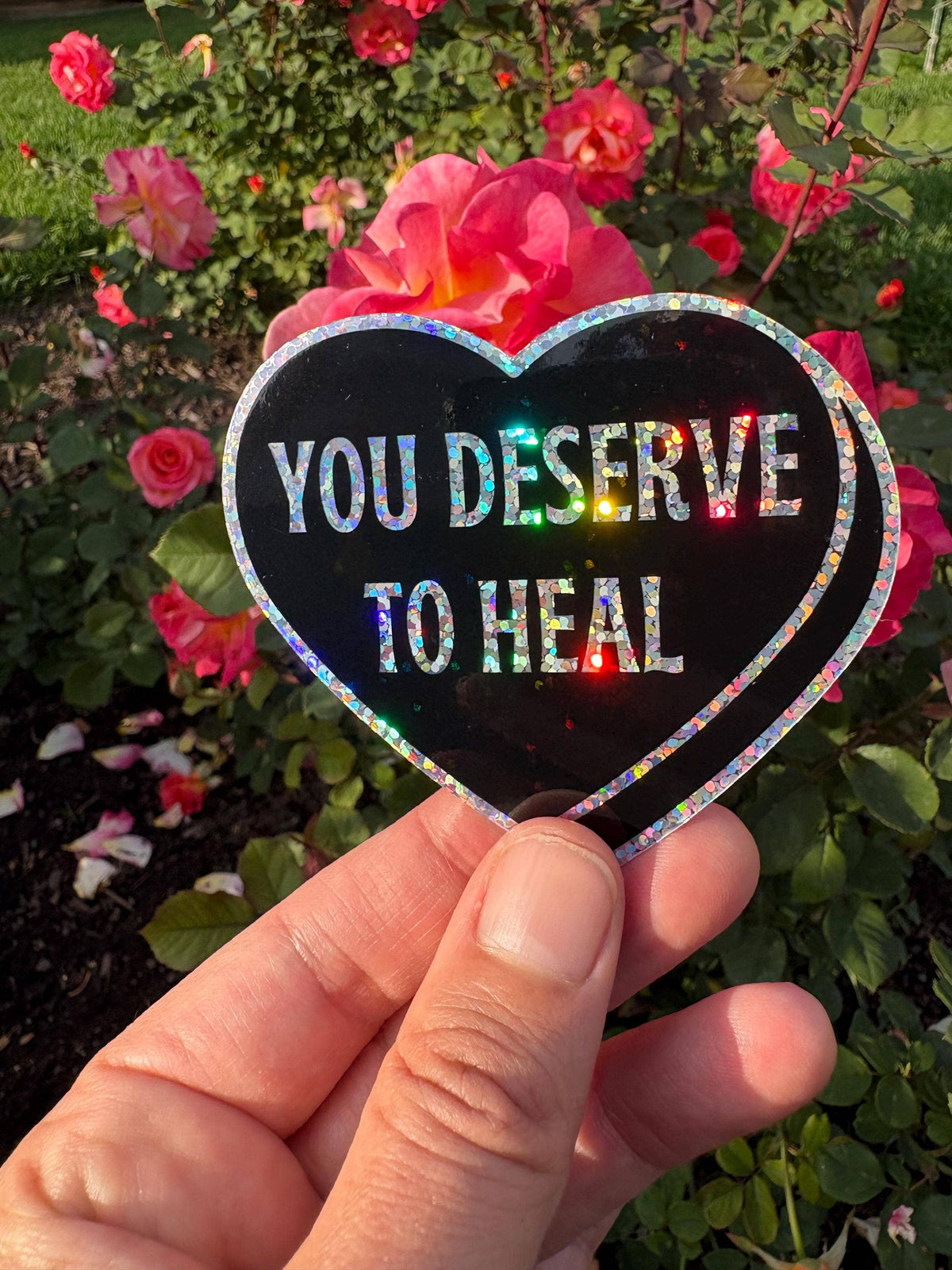 A hand holds a heart-shaped black sticker with holographic glitter edging and the text 'YOU DESERVE TO HEAL' in white, set against a background of pink roses and green foliage.