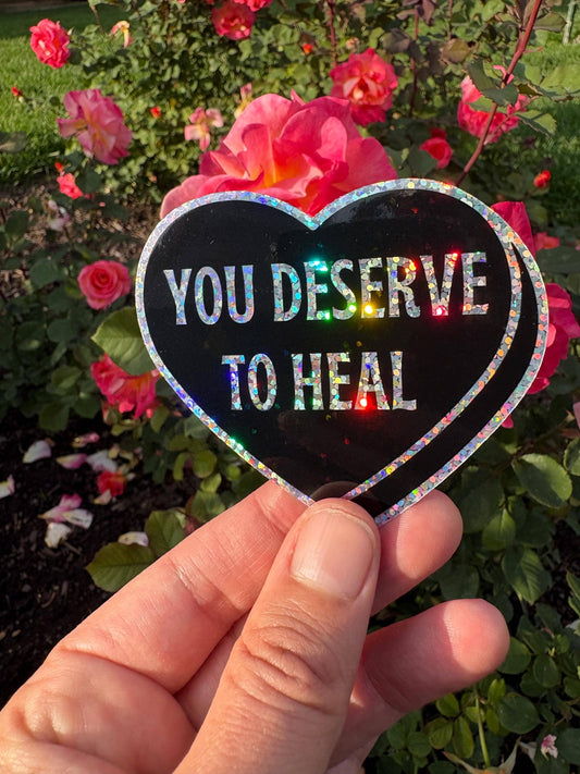 A hand holds a heart-shaped black sticker with holographic glitter edging and the text 'YOU DESERVE TO HEAL' in white, set against a background of pink roses and green foliage.