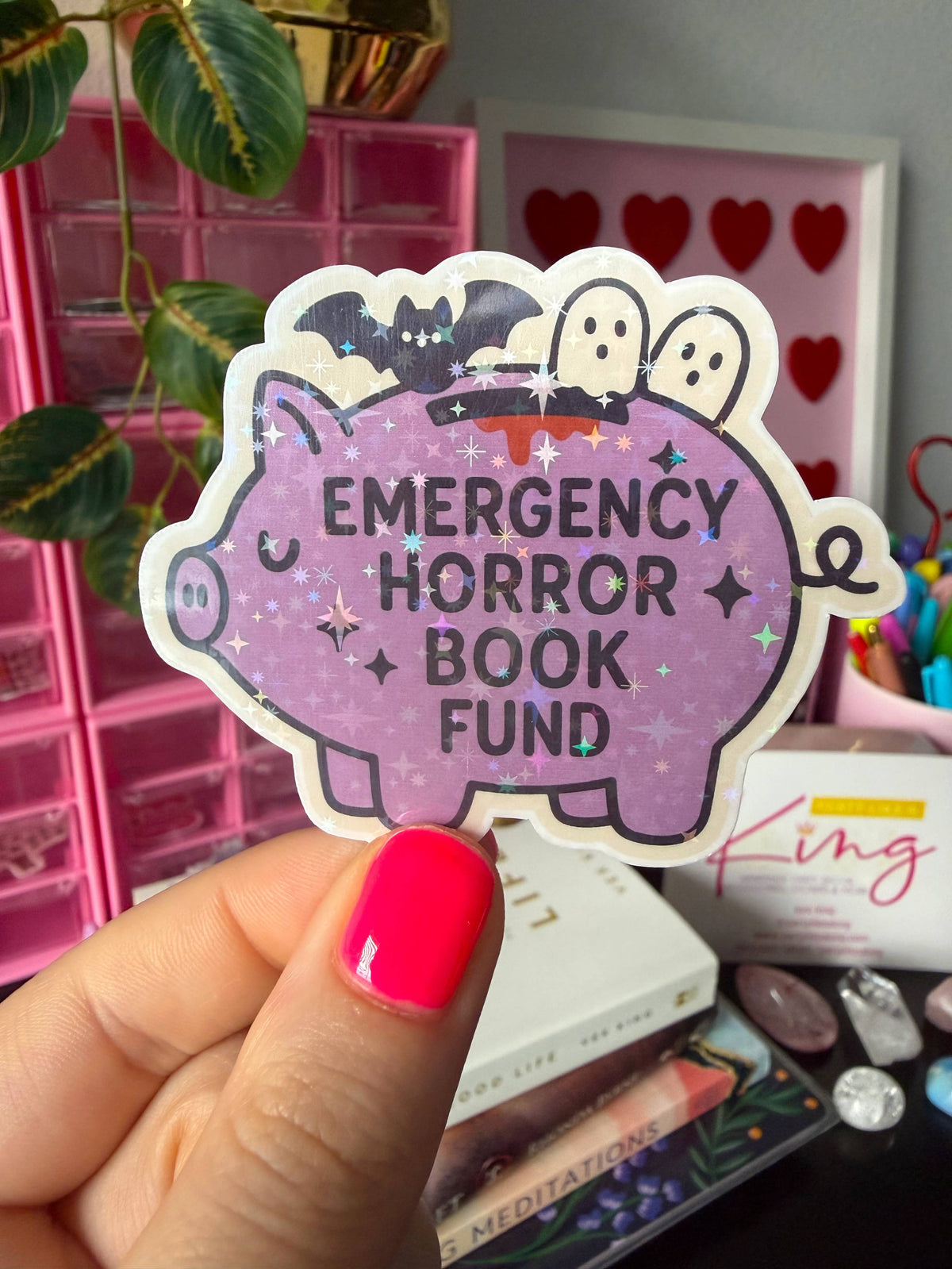 A hand with bright pink nail polish holds a die-cut sticker shaped like a purple pig. The sticker has black text reading 'EMERGENCY HORROR BOOK FUND' and Halloween-themed illustrations including bats, ghosts, and stars on a sparkly lavender background.