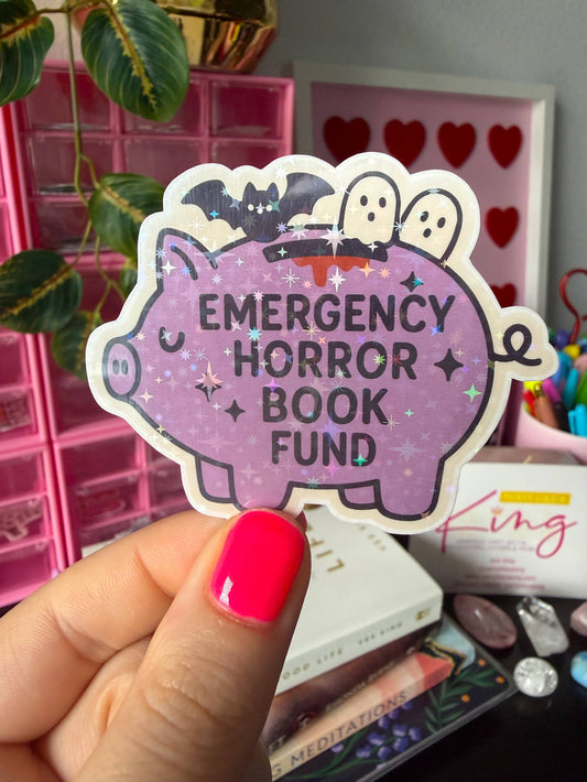 A hand with bright pink nail polish holds a die-cut sticker shaped like a purple pig. The sticker has black text reading 'EMERGENCY HORROR BOOK FUND' and Halloween-themed illustrations including bats, ghosts, and stars on a sparkly lavender background.