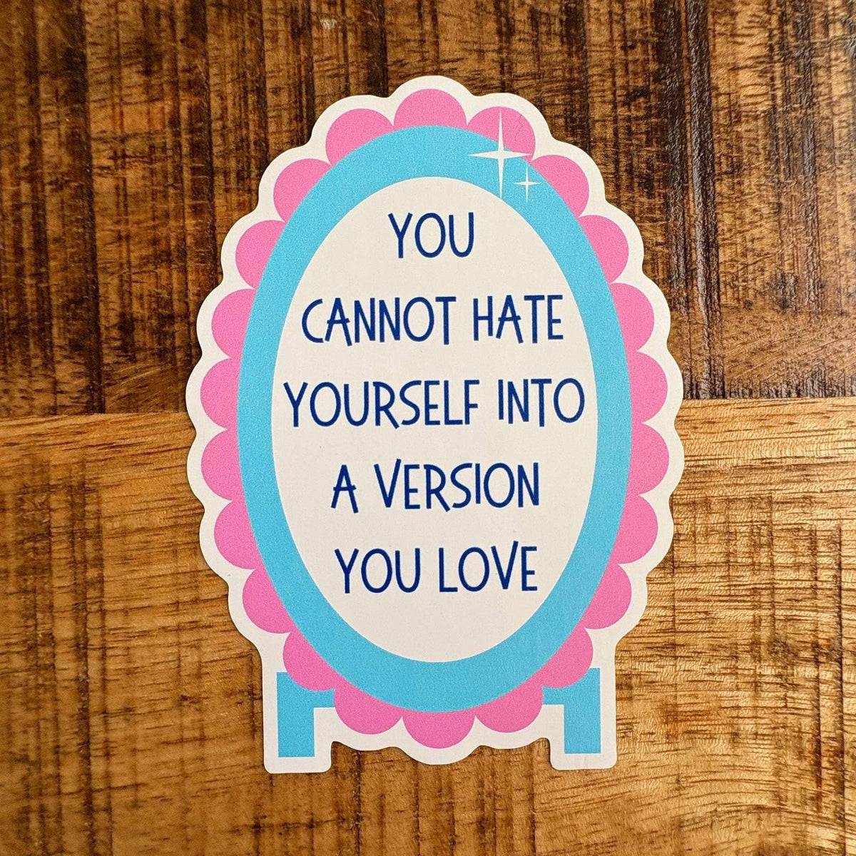 A die-cut sticker with the text 'YOU CANNOT HATE YOURSELF INTO A VERSION YOU LOVE' in blue lettering, featuring a white center, a light blue oval border, and a pink scalloped outer border, shown on a wooden surface.