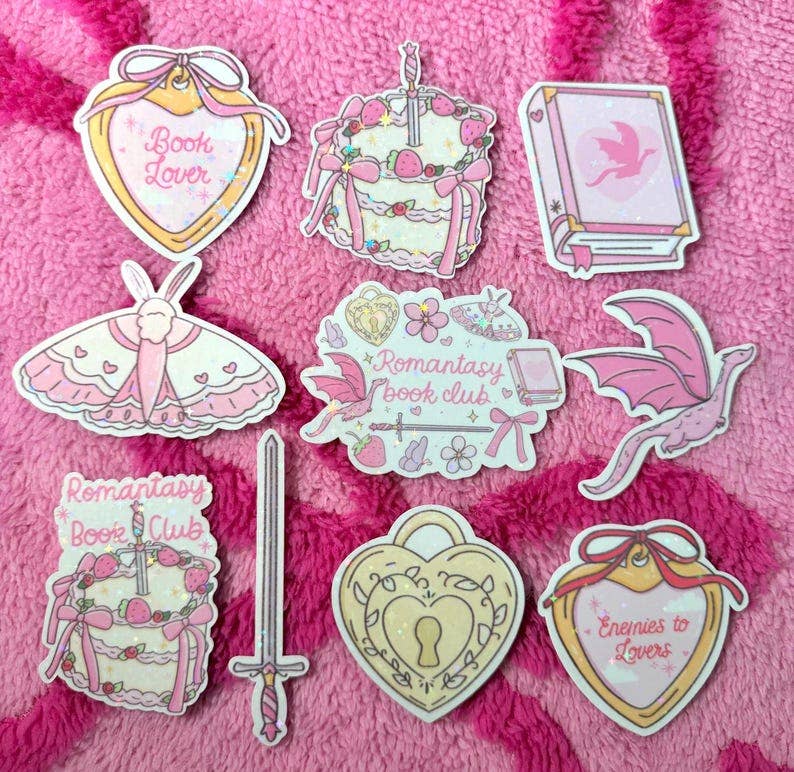 A set of cute, book-and-moth-themed enamel pins arranged on a pink textured fleece background. The pins feature illustrations of pink and cream-colored moths, a sword, a keyhole, and hearts with text such as 'Book Lover', 'Romantasy Book Club', and 'Enemies to Lovers'.