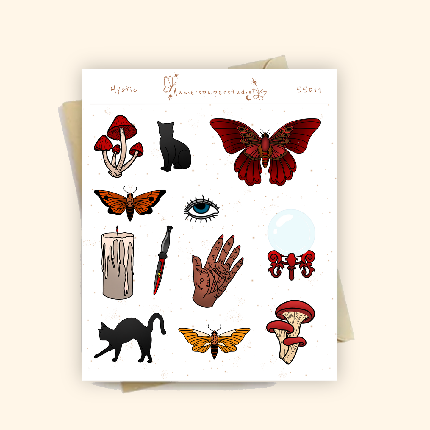A sheet of decorative stickers or decals with occult and mystical illustrations, including red mushrooms, a black cat, a red moth, an eye, a candle, a hand, and a dagger, shown on a white backing card over a beige envelope.