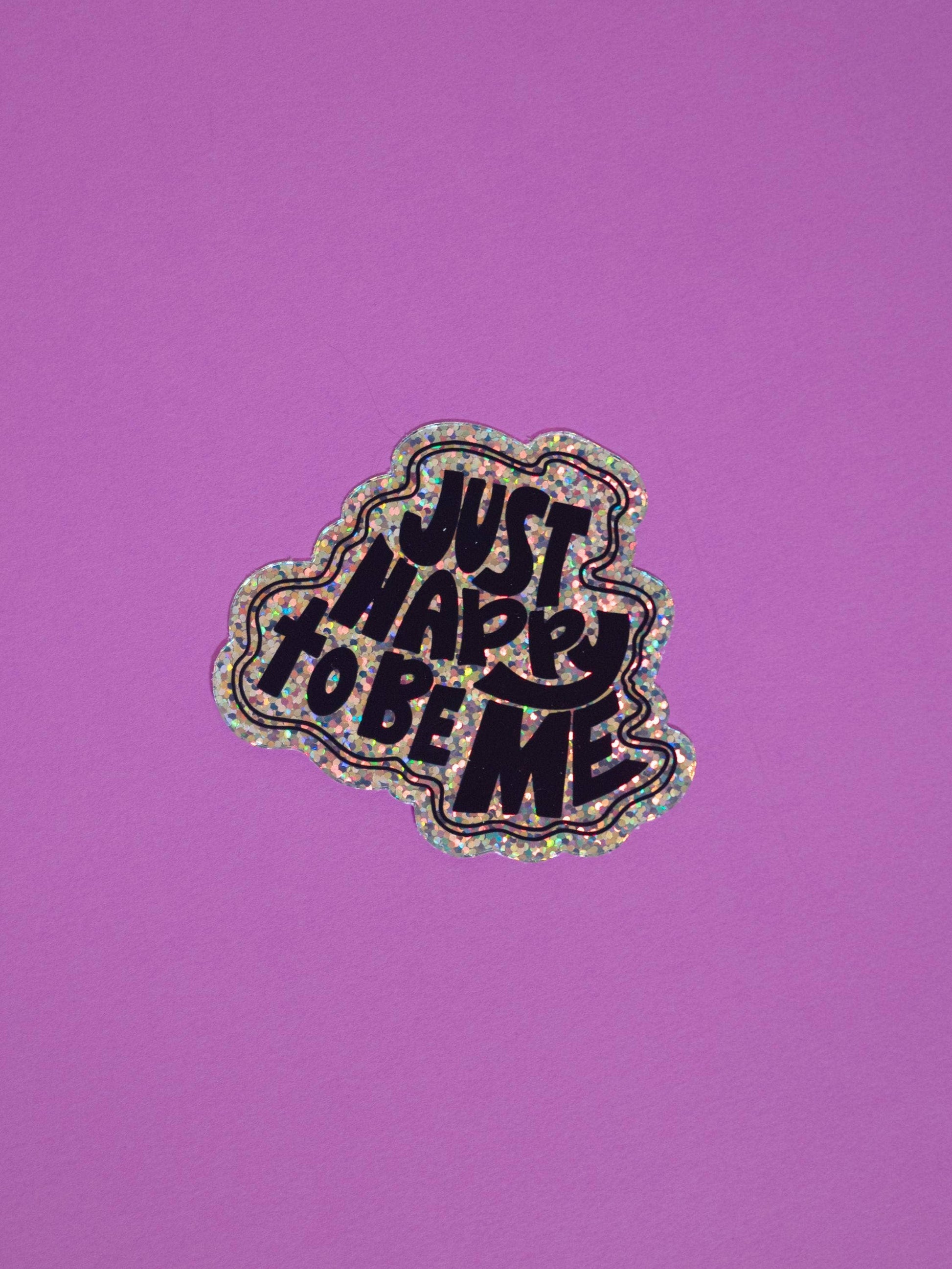 A die-cut sticker with the text 'JUST HAPPY TO BE ME' in black lettering, outlined in holographic glitter, shown on a solid purple background.
