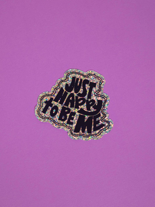 A die-cut sticker with the text 'JUST HAPPY TO BE ME' in black lettering, outlined in holographic glitter, shown on a solid purple background.