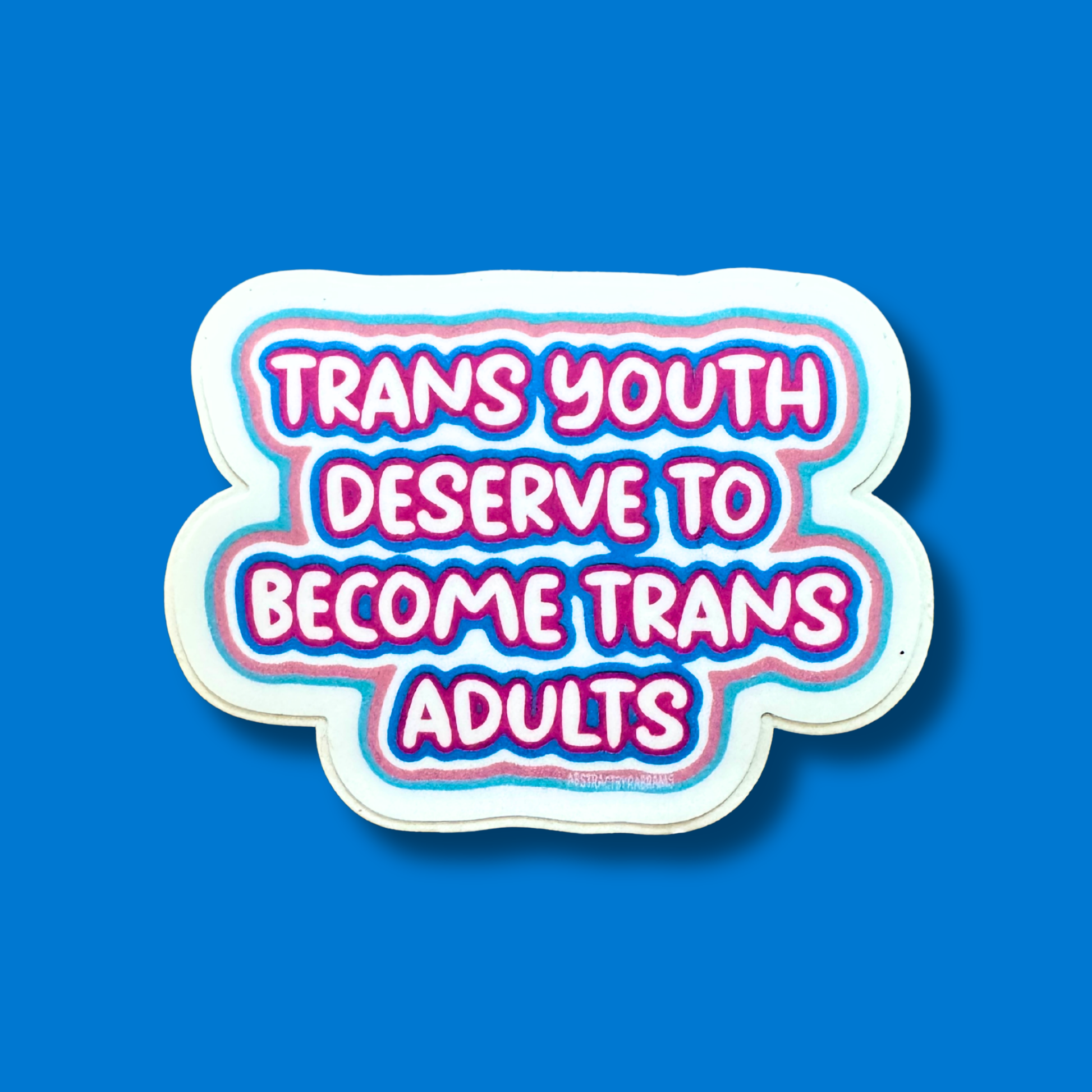 A die-cut sticker with the text 'TRANS YOUTH DESERVE TO BECOME TRANS ADULTS' in bold pink letters with blue and purple outlines, set against a solid blue background.
