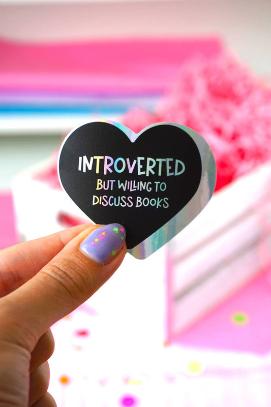 Heart-shaped sticker with 'Introverted but willing to discuss books' text held by a hand with colorful nail polish.