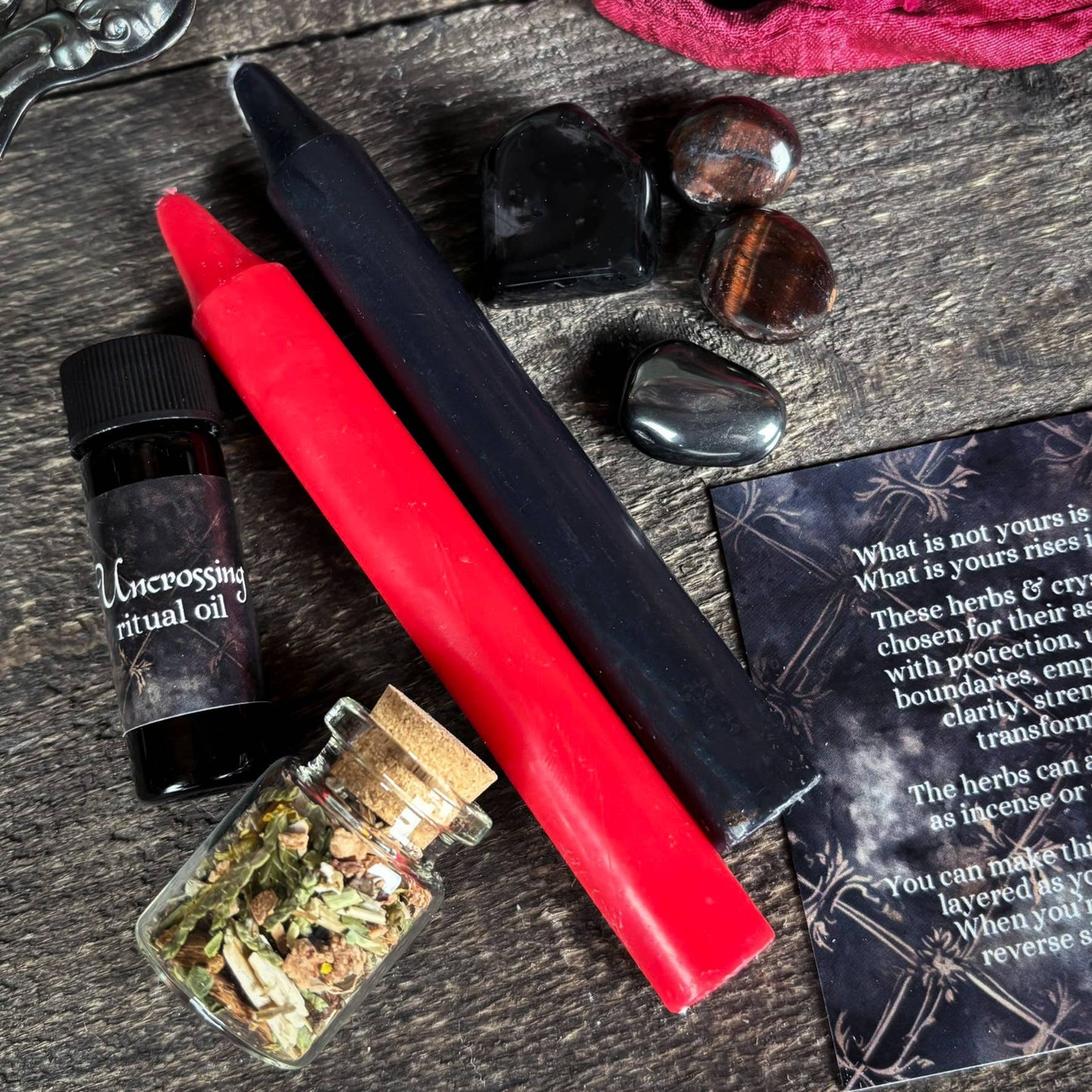 Red and black candles, herbs, and a bottle of oil on a wooden surface with text and stones.