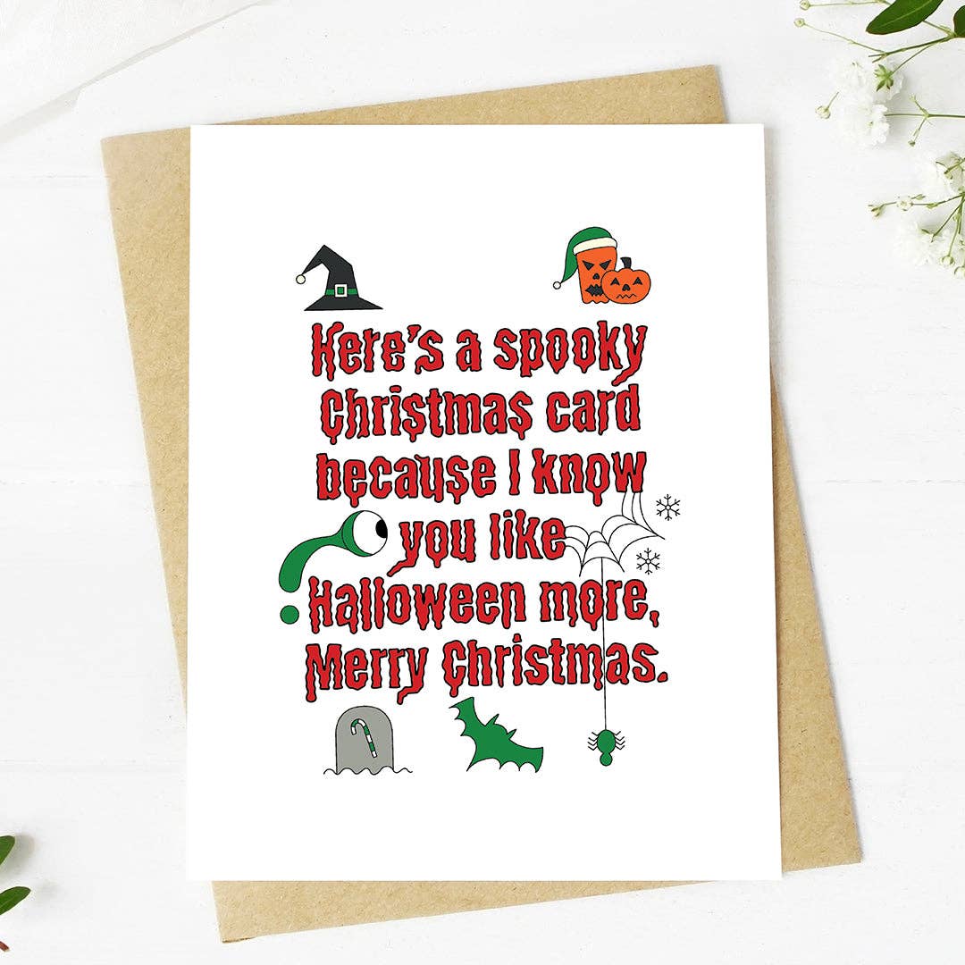 A white Christmas greeting card with red text reading 'Here's a spooky Christmas card because I know you like Halloween more, Merry Christmas.' surrounded by small Halloween illustrations, shown with a brown kraft envelope on a white surface.