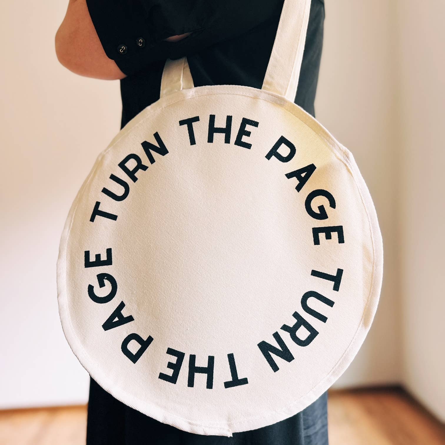 Round beige tote bag with 'TURN THE PAGE' text held by a person.