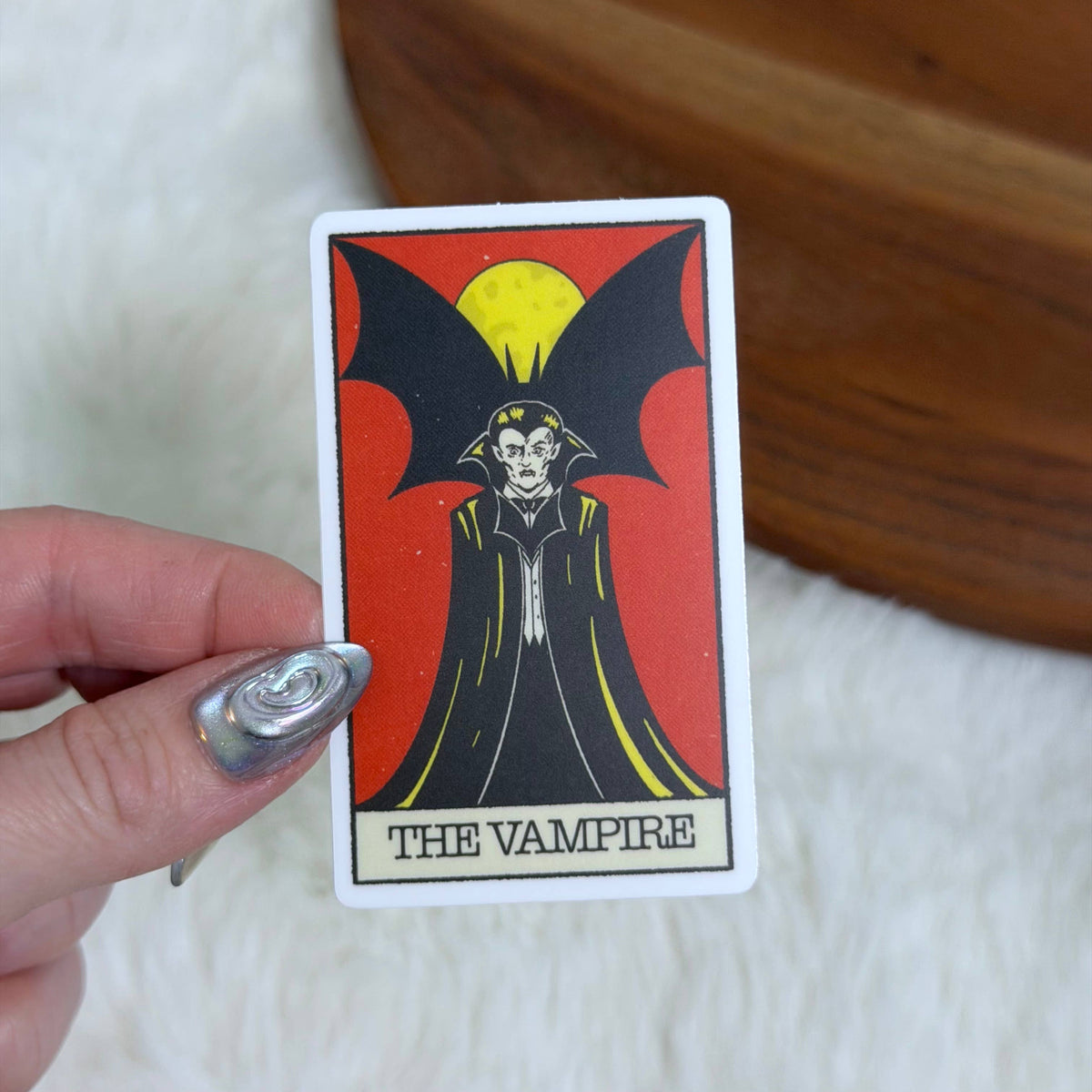 A hand with silver nail polish holds a tarot card featuring 'The Vampire' illustration against a red background. The card has a white border and is photographed over a white faux fur surface with a wooden surface in the background.