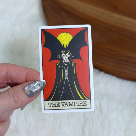 A hand with silver nail polish holds a tarot card featuring 'The Vampire' illustration against a red background. The card has a white border and is photographed over a white faux fur surface with a wooden surface in the background.