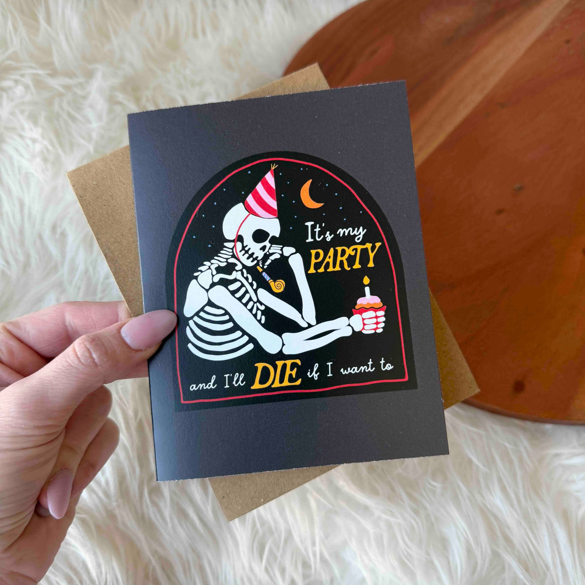 Greeting card with skeleton design and text held by a hand on a soft surface.