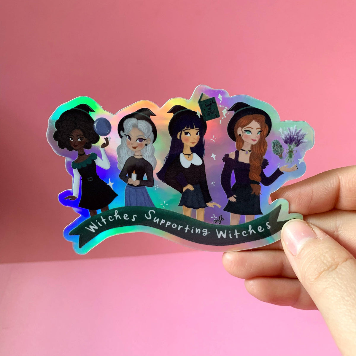 A hand holding a die-cut holographic sticker featuring four cartoon witches in black outfits with the text 'Witches Supporting Witches' on a pink background.