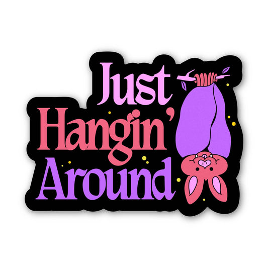 A die-cut sticker with the phrase 'Just Hangin' Around' in pink and purple lettering, featuring a cute cartoon bat hanging upside down on the right side, all on a black background with small yellow speckles.