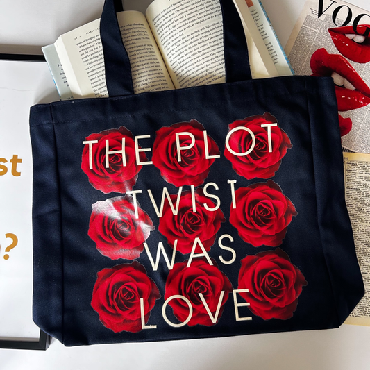 Black tote bag with red roses and 'The Plot Twist Was Love' text on a surface with books and a magazine.