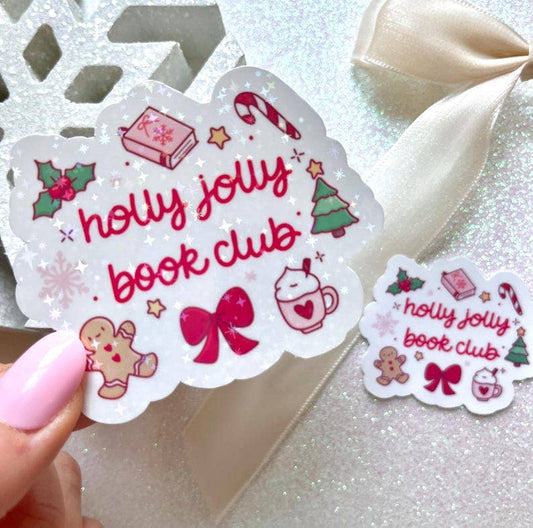 A hand with pink nail polish holds a scalloped white sticker that reads 'holly jolly book club' in red script, decorated with Christmas illustrations like a candy cane, holly, gingerbread man, bow, tree, and hot chocolate. A smaller matching sticker sits on glittery white paper beside a cream ribbon.