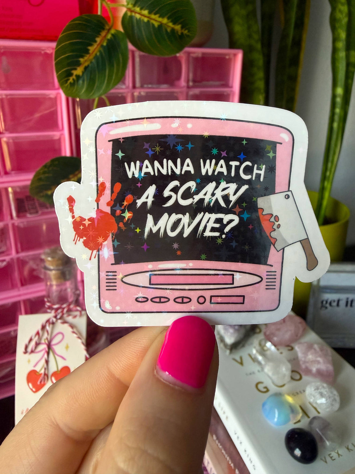 A hand with bright pink nail polish holds a die-cut sticker shaped like a pink retro television. The screen of the TV is black with the white text 'WANNA WATCH A SCARY MOVIE?' and colorful star accents.