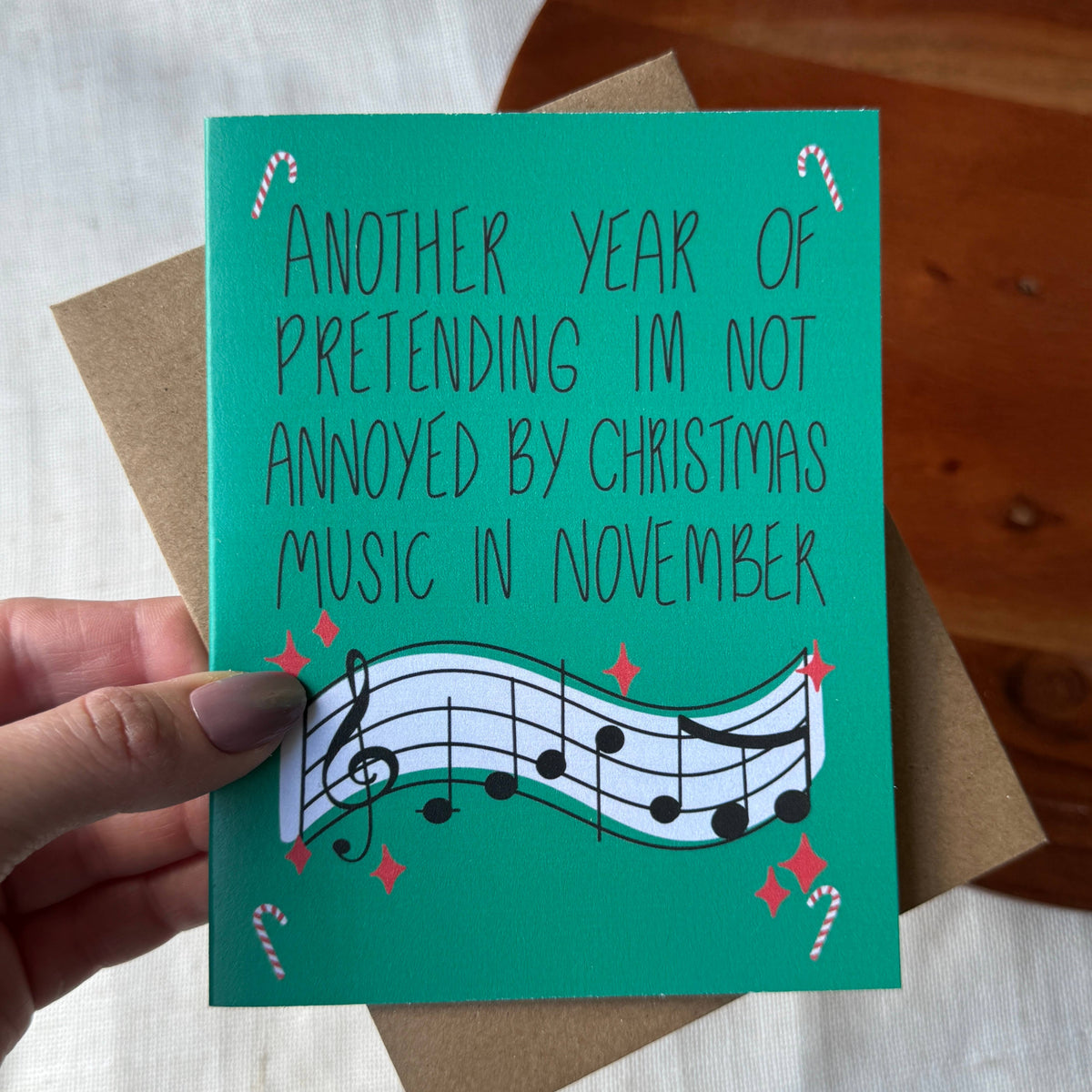 A hand holds a teal Christmas greeting card with the text 'ANOTHER YEAR OF PRETENDING IM NOT ANNOYED BY CHRISTMAS MUSIC IN NOVEMBER' above a wavy musical staff. The card is shown with a brown envelope on a light fabric surface with a wooden table in the background.