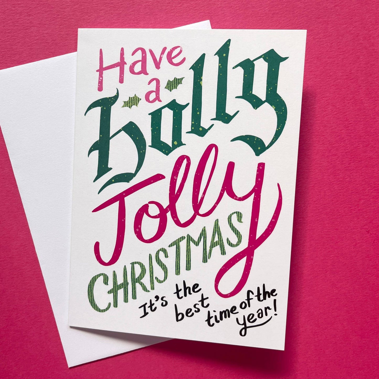 A white Christmas greeting card with festive hand-lettered text reading 'Have a Holly Jolly Christmas It's the best time of the year!' placed on top of a white envelope against a bright pink background.