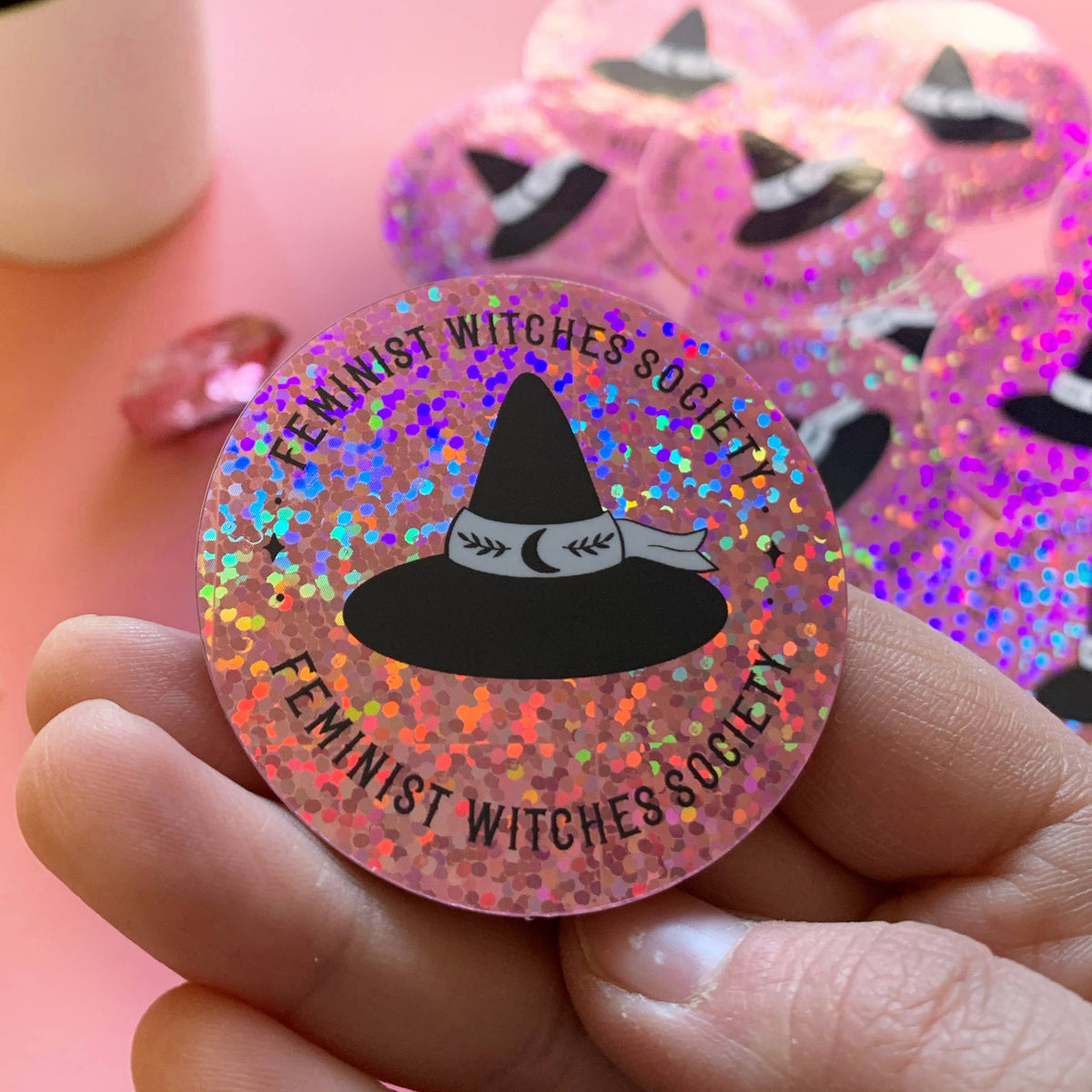 A hand holds a round glittery pink pinback button featuring a black witch hat graphic and the text 'FEMINIST WITCHES SOCIETY'. More matching pink glitter pins are blurred in the background.